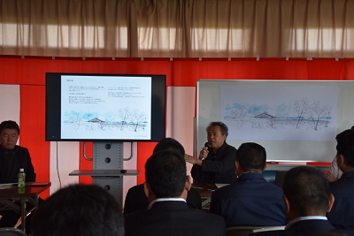 モニター横に立ち、マイクを使い話す男性の写真