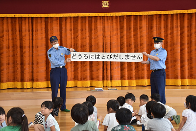 「どうろにはとびだしません」と書かれた紙の両端を持ち、園児たちに見せる警察官2人の写真