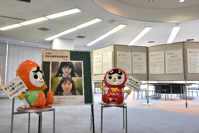 ポスターの貼られたパネルの両脇の椅子の上に赤とオレンジのキャラクターが座っている写真