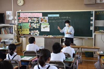 食べ物の写真や説明が書かれた黒板の前で授業を行う女性の写真