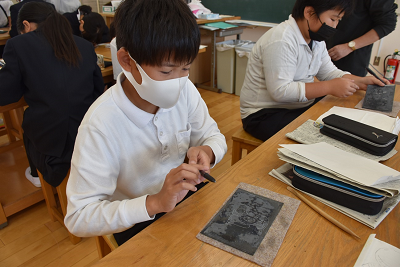 筆箱や銅板の置かれた机で男子児童がペンをもって作業している写真