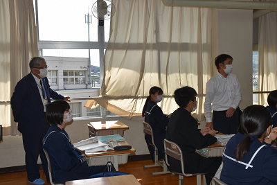 教室で立って話す男子学生を見るスーツ姿の男性の写真