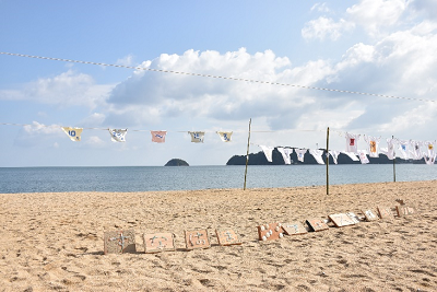 「すなはまフェスティバル」と書かれたボードとTシャツが飾られた浜辺の写真