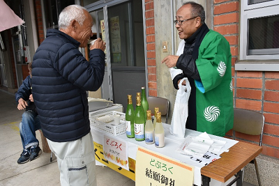 緑色の法被を着た男性と飲みながら話す男性の写真