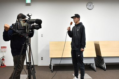 カメラの前でマイクを持ちながら話す男性の写真