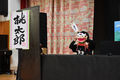 演目台に「桃太郎」と書かれたステージで鉢巻をした少年の人形が登場している写真