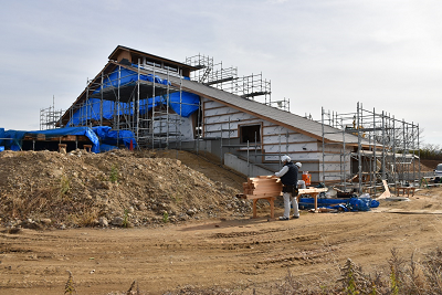 小さい坂と平面に建てられている建設中の建物に工事用の足場やブルーシートが掛けられている写真
