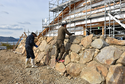 ヘルメットを着用した2人が石垣に登りながら建物を確認している写真
