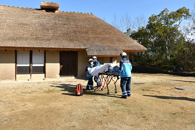 茅葺き屋根の古民家から担架が運び出されている写真