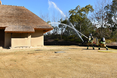 2名でホースを持ち、茅葺き屋根の古民家に向かって放水する消防団員の写真