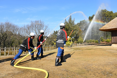 法被姿の消防団員3人が古民家に向けてホースで放水し、水しぶきに虹がかかっている写真