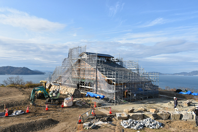 足場に覆われた建物の周りを重機や作業員が工事している写真