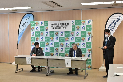 市のロゴ入りボードの前に長机が設置され、スーツ姿の男性2名が着席して話している写真