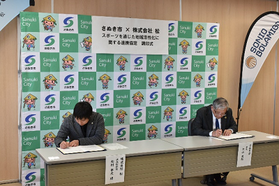 市のロゴ入りボードの前で書類にサインするスーツ姿の男性2人の写真