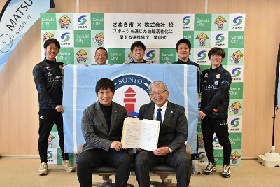 スーツ姿の2人が協定書を手に持ち、その後ろにユニフォーム姿の選手が並んでいる写真