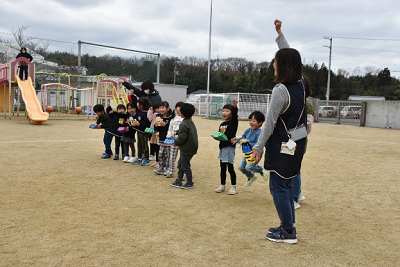 片腕をあげている女性の横で一列に並ぶ園児たちの写真