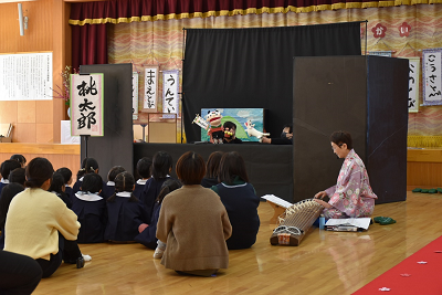 琴を弾く女性の隣でパペット人形劇を鑑賞する園児たちの写真