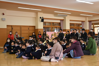 パイプ椅子に座る人たちの前で体育座りしている園児の写真