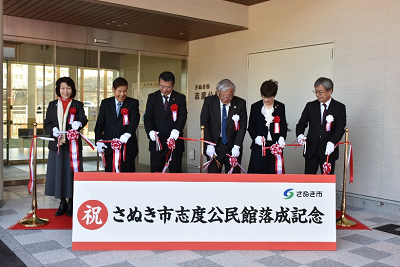 胸元にバラのコサージュを付けたスーツ姿の男女が赤白のテープを切っている写真