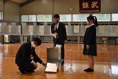 マイクを持って話す人と投票箱を開けて中を見せている男性の写真