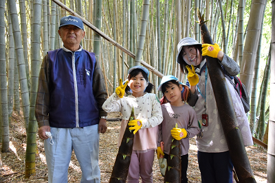 竹藪の中でタケノコをもってピースサインする園児と大人の写真