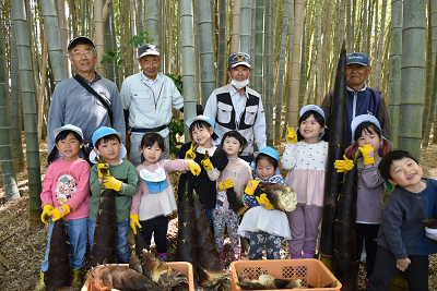 作業服を着た男性4人の前でタケノコをもって並ぶ園児たちの写真