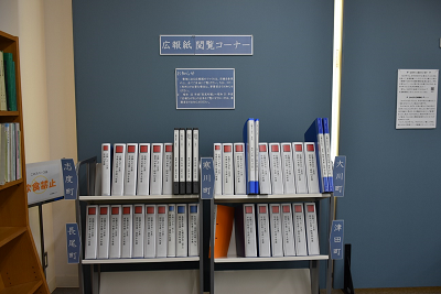 「広報紙閲覧コーナー」と書かれた壁の下に置かれた棚にファイルが陳列されている写真
