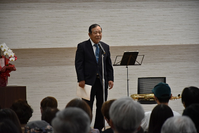 スーツ姿の男性がスタンドマイクを使って話している写真