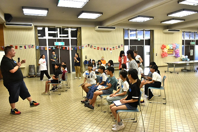 椅子に座る児童に向き合ってマイクを持って話す男性の写真