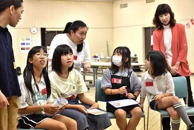 椅子に並んで座る女子児童4人と立ったまま話す女性の写真