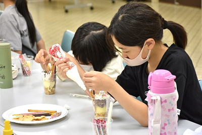 お菓子などが入った容器に生クリームを絞る女子児童の写真