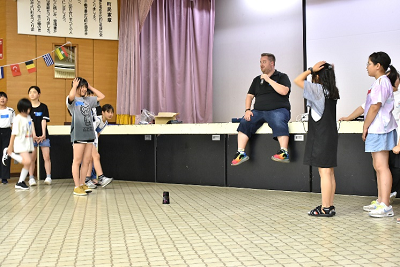 床に置かれた黒いカップを中心に向かい合って立つ女子児童が頭に手を乗せている写真