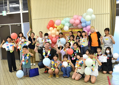 風船の前で首飾りをつけてポーズを決める児童たちと教師の写真