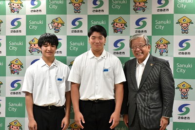 市のロゴ入りボードの前で半袖のワイシャツを着用した2人の生徒とスーツ姿の男性が並んでいる写真