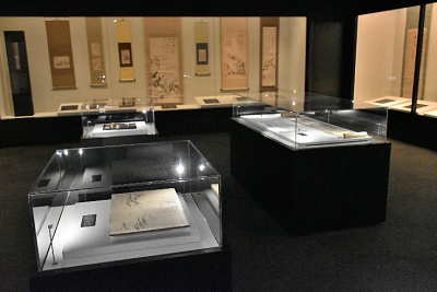 薄暗い展示室で、ガラスケースに古い書画や巻物が並び周囲の壁にも掛け軸が展示されている写真