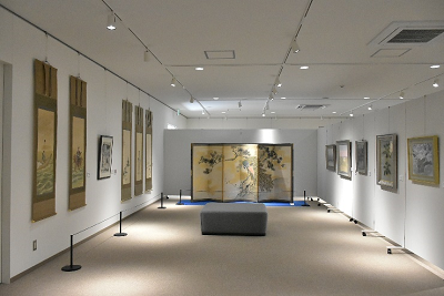 白い壁の展示室で、左右の壁に掛け軸や絵画が並び正面に屏風が置かれた写真