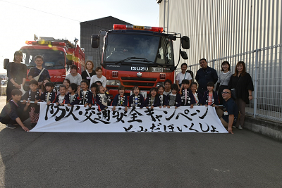 消防車の前に並んだ子どもたちや大人たちが白い横断幕を持っている写真
