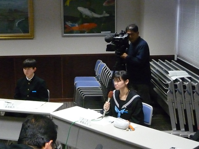 マイクを持った生徒の後ろにカメラを抱えた人が立っている写真