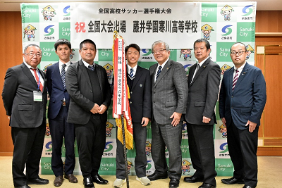 旗を持った生徒やスーツ姿の男性達が並んでいる写真