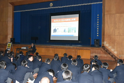 壇上に展開されたスクリーンのスライドを見る生徒達の写真