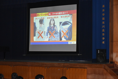 壇上のスクリーンに歩いている時、自転車の運転時、車の運転時の写真にバツ印が付けられたスライドが映されている写真