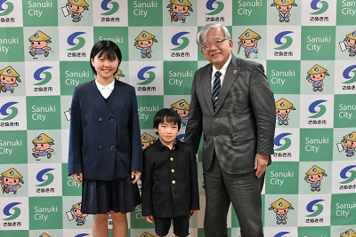 市のロゴ入りボードの前で学生服を着た2人の児童とスーツ姿の男性が並んでいる写真