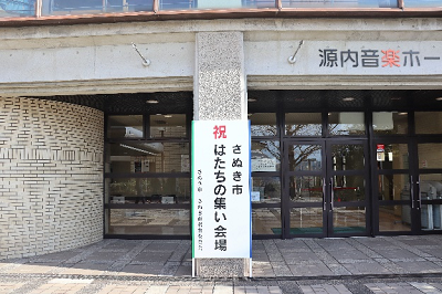「さぬき市 はたちの集い会場」と書かれた看板が建物入口に設置されている写真
