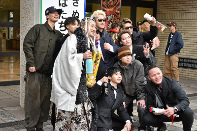 酒や花を手に看板の前でポーズする袴やスーツを着た男性たちの写真