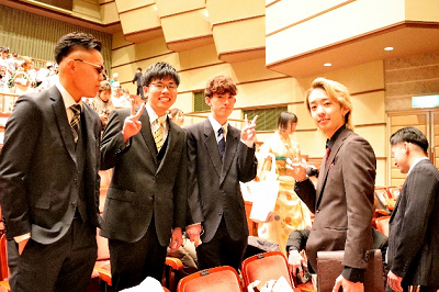 観客席で立ってピースするスーツを着た男性たちの写真