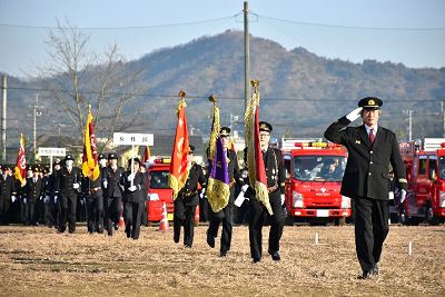 敬礼した男性に続いて、旗を持って行進する消防団制服姿の人たちの写真