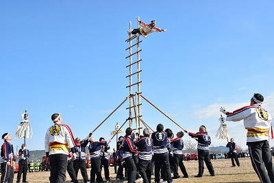 大勢の人が支える梯子の上で、地面と水平の姿勢になり両手を広げる人の写真