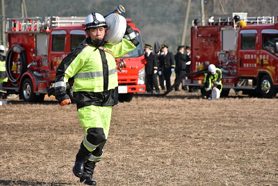 ヘルメットと防火服を着用した男性が左肩にホースを担いで走っている写真