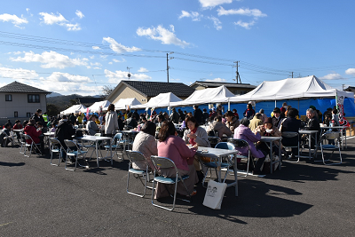 外にある白いテント前の机とパイプ椅子に座る人達の写真