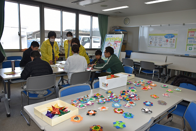 折り紙作品が置かれた机の奥で折り紙を折っている人達の写真